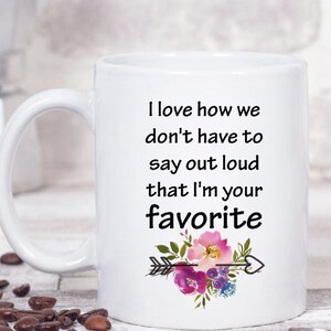 May include: White ceramic coffee mug with a black text saying "I love how we don't have to say out loud that I'm your favorite" with a watercolor floral design and an arrow with a heart.