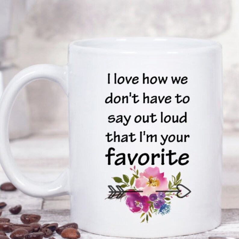 May include: White ceramic coffee mug with a black text saying "I love how we don't have to say out loud that I'm your favorite" with a watercolor floral design and an arrow with a heart.
