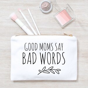 Puede incluir: Bolsa de maquillaje de lona blanca con cremallera dorada. La bolsa tiene texto negro que dice "Good Moms Say Bad Words" con un peque&ntilde;o dise&ntilde;o floral negro debajo del texto.