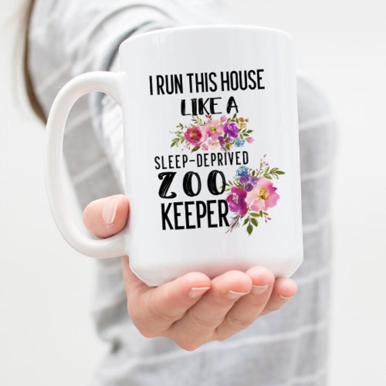 Może przedstawiać: Biały kubek ceramiczny z czarno-białym nadrukiem, na kt&oacute;rym widnieje napis "Prowadzę ten dom jak pozbawiony snu opiekun zoo". Nadruk otoczony jest r&oacute;żowymi, fioletowymi i niebieskimi kwiatami akwarelowymi.