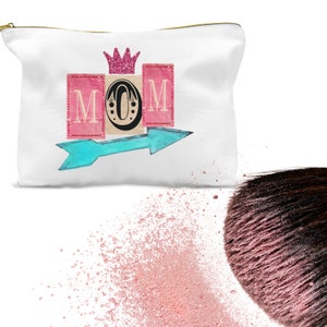 K&ouml;nnte beinhalten: Wei&szlig;e Kosmetiktasche aus Canvas mit einem rosa, blauen und goldenen Design. Die Tasche zeigt das Wort "MOM" in rosa und blauen Bl&ouml;cken mit einer goldenen Krone &uuml;ber den Buchstaben. Ein blauer Pfeil zeigt nach rechts.