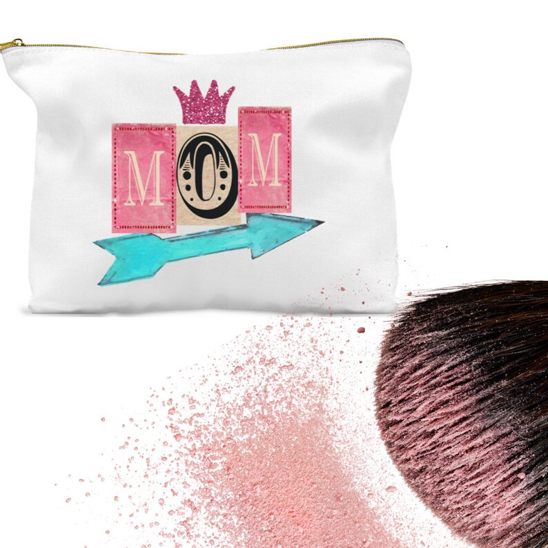 K&ouml;nnte beinhalten: Wei&szlig;e Kosmetiktasche aus Canvas mit einem rosa, blauen und goldenen Design. Die Tasche zeigt das Wort "MOM" in rosa und blauen Bl&ouml;cken mit einer goldenen Krone &uuml;ber den Buchstaben. Ein blauer Pfeil zeigt nach rechts.