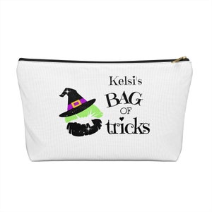 May include: White zippered pouch with a black zipper. The pouch has a green witch's hat with a purple band and a black witch's mouth with glitter. The text on the pouch reads "Kelsi's Bag of tricks".