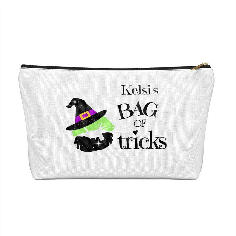 May include: White zippered pouch with a black zipper. The pouch has a green witch's hat with a purple band and a black witch's mouth with glitter. The text on the pouch reads "Kelsi's Bag of tricks".