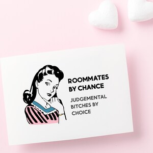May include: A white card with a black and white illustration of a woman with a pink and black striped shirt. The text on the card reads "Roommates by chance, Judgemental Bitches by choice."