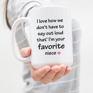 May include: White ceramic coffee mug with a black handle. The mug has a black text message that reads "I love how we don't have to say out loud that I'm your favorite niece" with a pink heart.