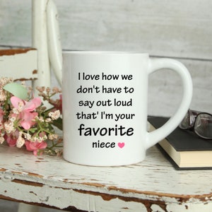 May include: White ceramic coffee mug with a black inscription that reads "I love how we don't have to say out loud that I'm your favorite niece". A pink heart symbol is at the end of the text.