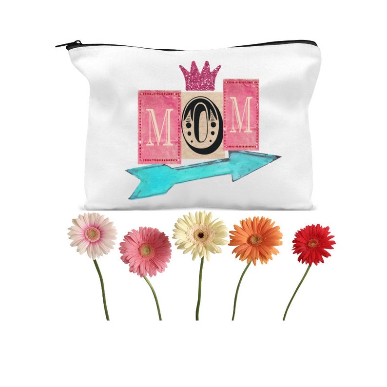 K&ouml;nnte beinhalten: Eine wei&szlig;e Rei&szlig;verschlusstasche mit einer rosa Krone und dem Wort "MOM" in rosa und blauen Buchstaben mit einem t&uuml;rkisfarbenen Pfeil, der nach rechts zeigt. F&uuml;nf rosa, orange und wei&szlig;e Gerbera-G&auml;nsebl&uuml;mchen sind in einer Reihe unter der Tasche angeordnet.