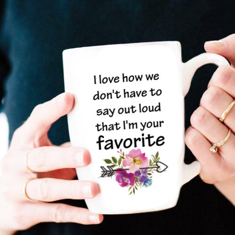 May include: White ceramic mug with a black and white quote that reads "I love how we don't have to say out loud that I'm your favorite". The quote is accompanied by a pink and purple floral design with an arrow.