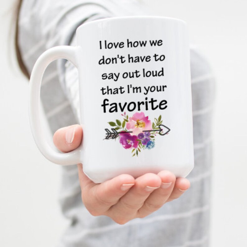 May include: White ceramic mug with a black handle and a floral design. The mug has the text "I love how we don't have to say out loud that I'm your favorite" printed on it.