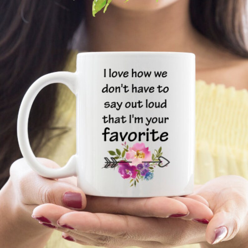 May include: White ceramic mug with a black and white quote that reads "I love how we don't have to say out loud that I'm your favorite". The quote is surrounded by a floral design with pink, purple, and green flowers and a black arrow.