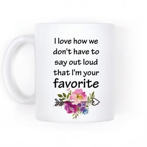 May include: White ceramic mug with a black and white quote that reads "I love how we don't have to say out loud that I'm your favorite". The quote is surrounded by a floral design with pink, purple, and blue flowers and a black arrow with a heart.