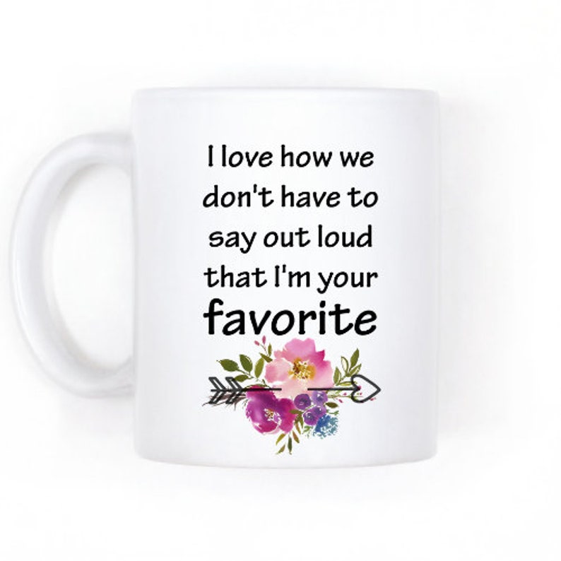 May include: White ceramic mug with a black and white quote that reads "I love how we don't have to say out loud that I'm your favorite". The quote is surrounded by a floral design with pink, purple, and blue flowers and a black arrow with a heart.