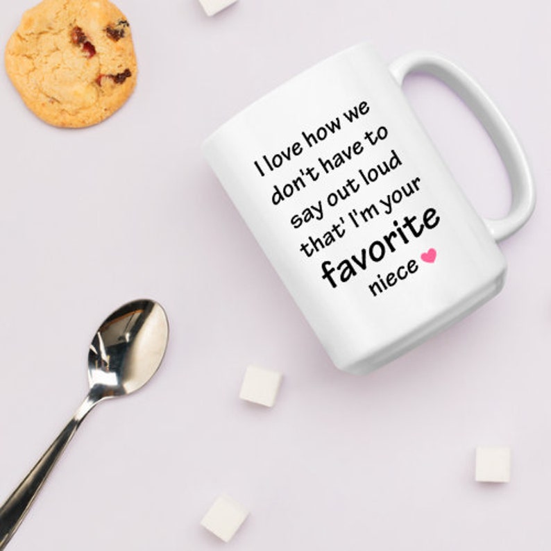 May include: White ceramic coffee mug with black text that reads "I love how we don't have to say out loud that I'm your favorite niece". A pink heart symbol is next to the word "niece".