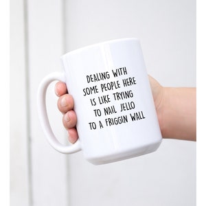 Może przedstawiać: Biały ceramiczny kubek z dużym uchwytem, trzymany w dłoni. Na kubku znajduje się czarny tekst o treści: "DEALING WITH SOME PEOPLE HERE IS LIKE TRYING TO NAIL JELLO TO A FRIGGIN WALL."