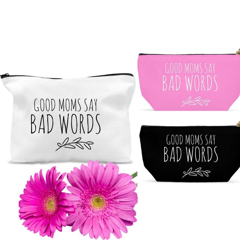 Puede incluir: Tres bolsas de maquillaje con el texto "Good Moms Say Bad Words" en letras negras sobre fondo blanco, rosa y negro. Las bolsas est&aacute;n rodeadas de dos margaritas rosas.