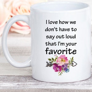 May include: White ceramic mug with a black and white quote that reads "I love how we don't have to say out loud that I'm your favorite". The quote is surrounded by a floral design with pink, purple, and green flowers and a black arrow.