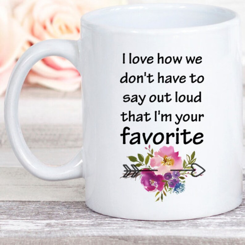 May include: White ceramic mug with a black and white quote that reads "I love how we don't have to say out loud that I'm your favorite". The quote is surrounded by a floral design with pink, purple, and green flowers and a black arrow.