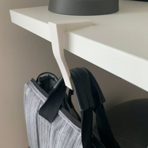 May include: A white, L-shaped desk hook is attached to the edge of a white desk. A gray and black bag hangs from the hook. The hook is designed to hold items, maximizing space and organization.