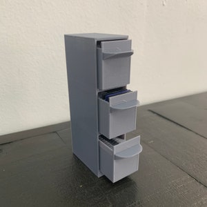 May include: A gray, three-drawer storage unit with pull-out drawers. The drawers are partially open, revealing the contents. The unit is made of a solid material and sits on a dark surface. The design is simple and functional.