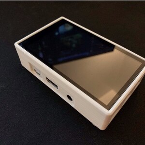 May include: A rectangular, cream-coloured electronic device with a black screen. The device has a sleek design with a black border around the screen. It features ports on the side, including a USB port and a headphone jack.