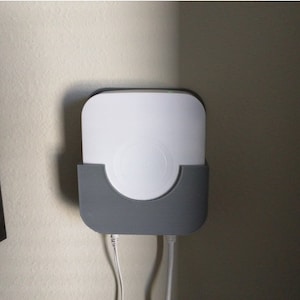 May include: A white, rectangular device with rounded corners is mounted on a wall. It is held in place by a gray, curved holder. Two white cords extend from the bottom of the device. The device is likely an electronic component.