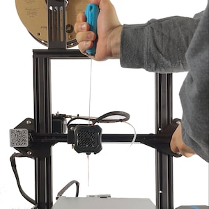 May include: A close-up of a 3D printer with a blue tool being used to feed filament into the extruder. The printer has a black frame and a white background.