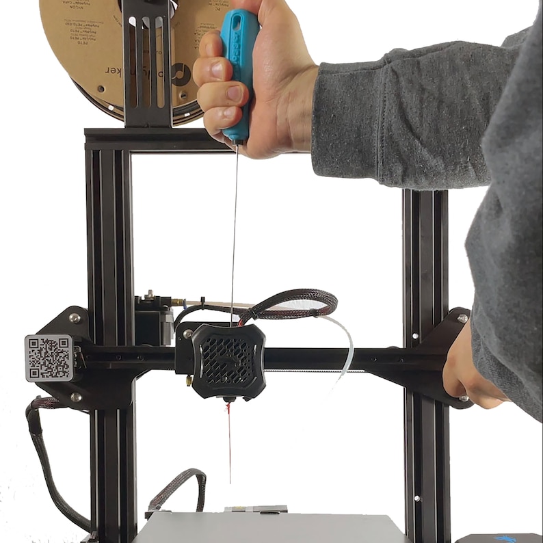 May include: A close-up of a 3D printer with a blue tool being used to feed filament into the extruder. The printer has a black frame and a white background.