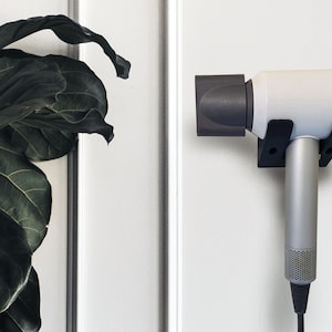 May include: A silver and white hair dryer is mounted on a black wall bracket. The hair dryer is plugged in and ready for use. A large green plant is visible on the left side of the image.