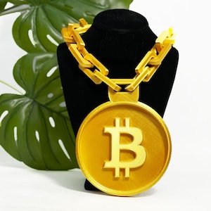 Könnte beinhalten: Eine goldfarbene Halskette mit einem großen Bitcoin-Anhänger. Der Anhänger zeigt das Bitcoin-Symbol. Die Halskette wird auf einem schwarzen Schaufensterkopf präsentiert, mit einem großen grünen Blatt im Hintergrund.