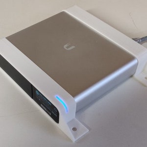 May include: A silver and black electronic device with a white frame and mounting brackets. The device has a small display with text and a blue light. A cable extends from the side.