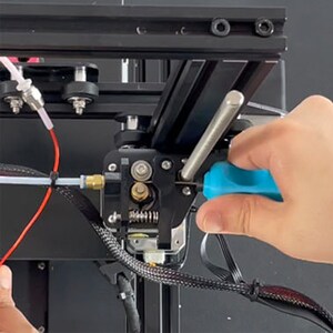 May include: A close-up of a 3D printer extruder with a blue screwdriver being used to tighten a screw. The extruder is made of black plastic and has a silver metal nozzle. There is a red wire and a clear tube connected to the extruder.