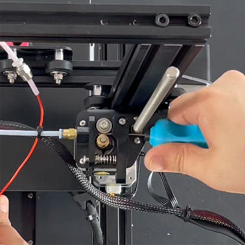 May include: A close-up of a 3D printer extruder with a blue screwdriver being used to tighten a screw. The extruder is made of black plastic and has a silver metal nozzle. There is a red wire and a clear tube connected to the extruder.