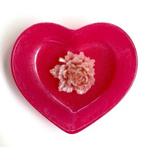 May include: A pink heart-shaped dish with a pink flower in the center. The dish is made of resin and has a glittery finish.