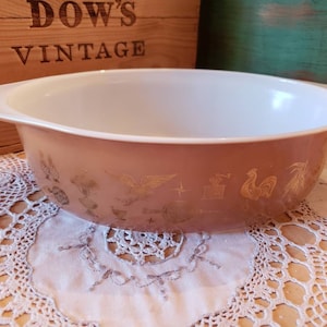 May include: A brown casserole dish with a white interior and a gold design of roosters, wheat, and other farm animals. The dish is sitting on a white crocheted doily.