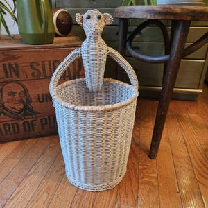 May include: A light gray wicker basket with a matching monkey figure attached. The basket has a round shape with a woven design and a light brown rim. The monkey figure is also made of wicker and has a detailed face.