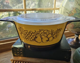May include: A vintage yellow Pyrex casserole dish with a clear lid. The dish features a brown fruit design and black handles. It is sitting on a green book and a small wooden stand. A globe is visible in the background.
