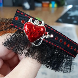 Puede incluir: Gargantilla de cinta negra con un colgante de corazón rojo con llamas, cuernos y cola. La gargantilla tiene detalles de pedrería roja y un ribete de encaje negro, creando una estética gótica.