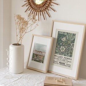 May include: Two framed prints on a white table with a white vase and dried flowers. The print on the left is a vintage image of a building. The print on the right is a floral design with the text "William Morris, Centenary Exhibition, Victoria & Albert Museum, Nearest Station South Kensington".