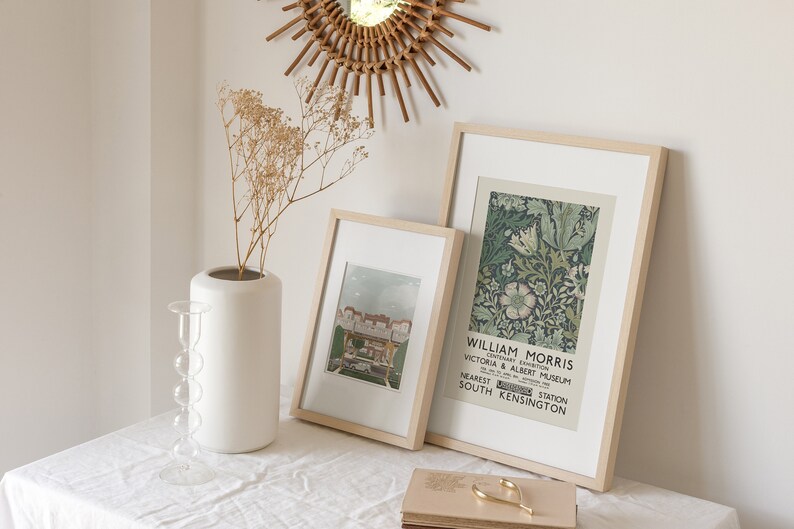 May include: Two framed prints on a white table with a white vase and dried flowers. The print on the left is a vintage image of a building. The print on the right is a floral design with the text "William Morris, Centenary Exhibition, Victoria & Albert Museum, Nearest Station South Kensington".