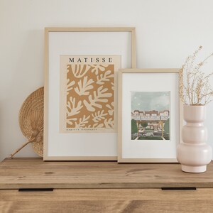 May include: Two framed prints on a wooden dresser. The print on the left is a Matisse print with the text "MATISSE papiers d&eacute;coup&eacute;s". The print on the right is a colorful illustration of a building with a blue sky.