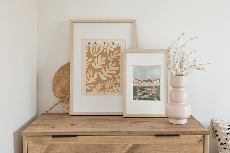 May include: Two framed prints on a wooden dresser. The print on the left is a Matisse print with the text "MATISSE papiers d&eacute;coup&eacute;s". The print on the right is a colorful illustration of a building with a blue sky.