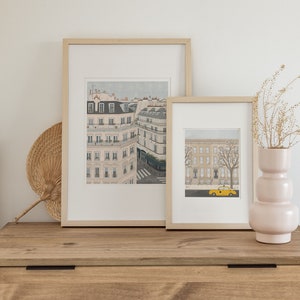 May include: Two framed illustrations of cityscapes in a light wood frame. The larger illustration shows a street scene with buildings and a crosswalk. The smaller illustration shows a street scene with buildings and a yellow taxi cab. The illustrations are on a wooden dresser with a wicker fan and a pink vase with dried flowers.