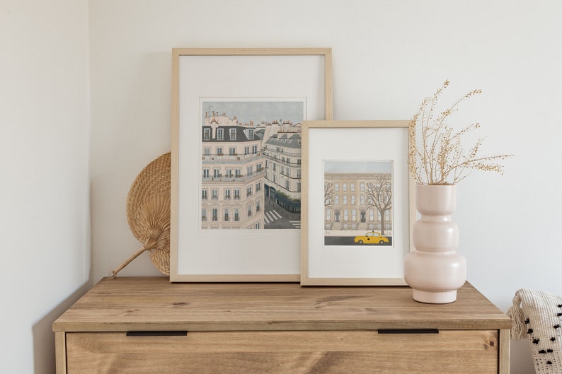 May include: Two framed illustrations of cityscapes in a light wood frame. The larger illustration shows a street scene with buildings and a crosswalk. The smaller illustration shows a street scene with buildings and a yellow taxi cab. The illustrations are on a wooden dresser with a wicker fan and a pink vase with dried flowers.