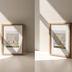 May include: Two framed illustrations of a city street scene with a yellow taxi cab in front of a brownstone building. The frames are made of natural wood and have a white mat.