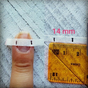 May include: A close-up of a finger with a white measuring tape wrapped around it. A yellow ruler is placed next to the finger, showing a measurement of 14 mm. The text "14 mm" is written in pink.