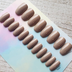 May include: A set of 10 nude-colored coffin-shaped press-on nails. The nails are arranged in two rows on a white and iridescent background.