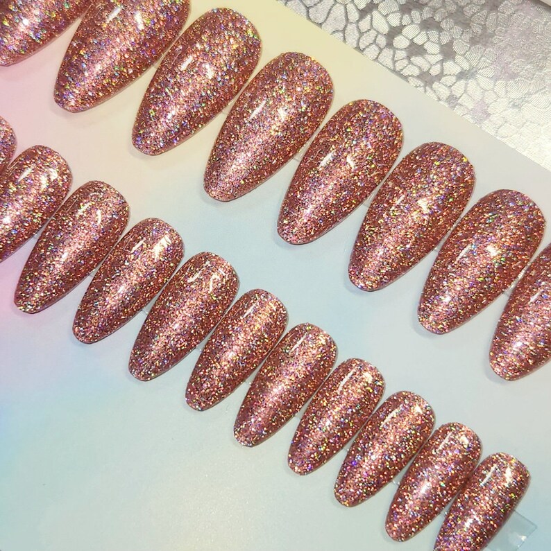 May include: A collection of rose gold glitter press-on nails. The nails are oval-shaped and covered in sparkling glitter. The light catches the glitter, creating a shimmering effect. The nails are arranged on a white surface.