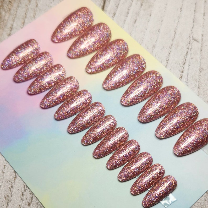 May include: A set of rose gold glitter press-on nails in various sizes. The nails are almond-shaped and have a sparkly, textured appearance. The glitter is evenly distributed across the surface of each nail, creating a shimmering effect. The nails are arranged on a colorful background.