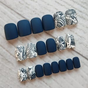 May include: A collection of navy blue matte press-on nails, some with intricate black and white floral and paisley designs. The nails are arranged in rows, showcasing various styles and patterns for a manicure.